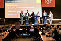 Das Foto zeigt die Teilnehmer*innen der Podiumsdiskussion. Außerdem sieht man eine Kollage, die die schwierige Situation auf dem Wohnungsmarkt symbolisiert. Dort werden diverse Wohnungsgesuche abgebildet.