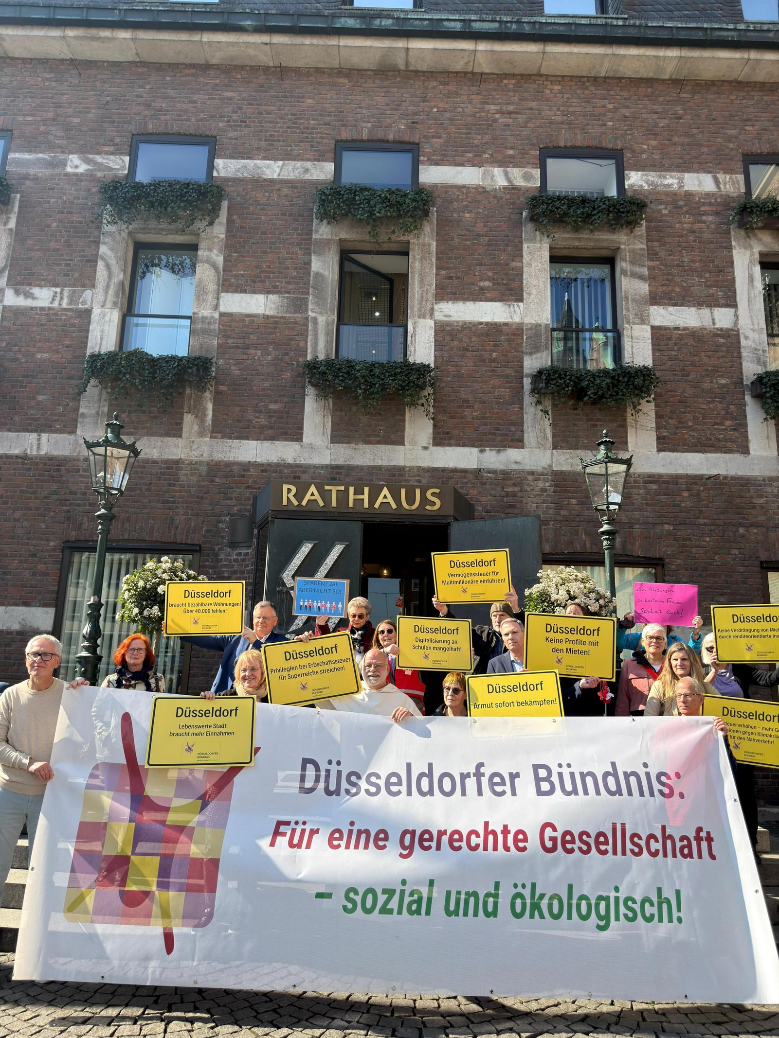 Die Bündnismitglieder stehen mit einem großen Transparent und vielen Schildern auf den Treppen des Düsseldorfer Rathauses. Sie machen auf ihre Forderungen gegenüber der Politik aufmerksam.
