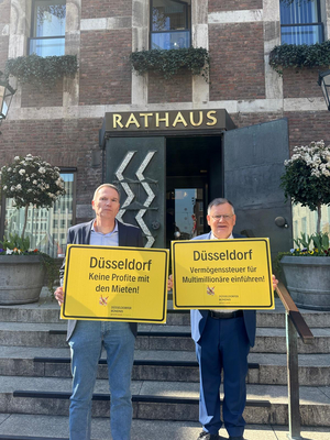 Claus Nesemann und Hans-Jochem Witzke stehen auf den Stufen des Düsseldorfer Rathauses und halten zwei Schilder mit den Aufschriften Düsseldorf- Keine Profite mit der Miete und Düsseldorf – Vermögenssteuer für Multimillionäre einführen! In ihren Händen.