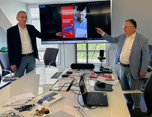 Auf dem Foto sind Claus Nesemann und Hans-Jochem Witzke zu sehen, wie sie auf einem großen Bildschirm zeigen. Auf dem Bildschirm sieht man ein Bild, auf dem für das neue Tool „Mietwucher melden!“ geworben wird.